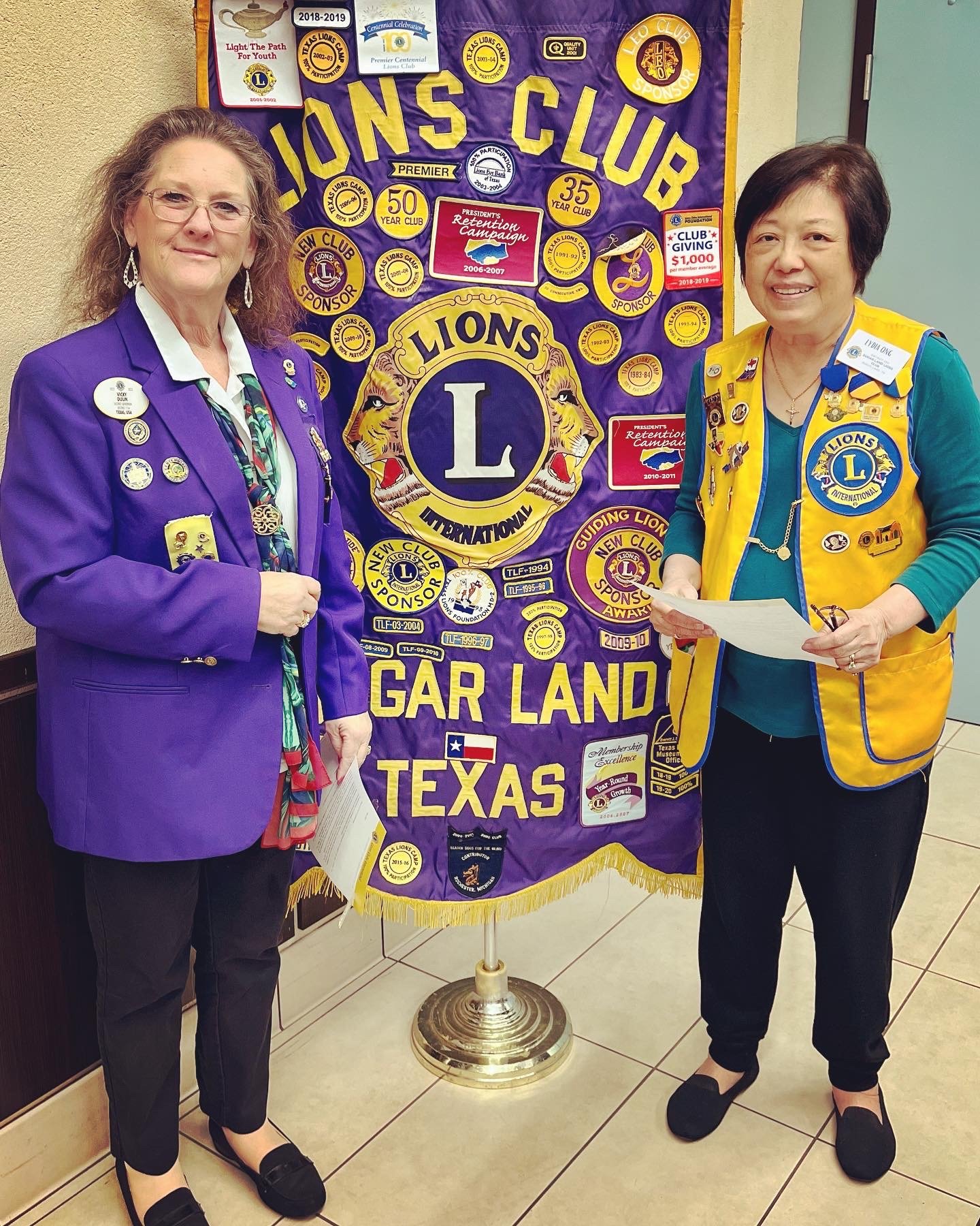 District Gov Vicki Dulin presents Lion Lydia with 10 year Chevron award in front of the Sugar Land Lions Club Banner.