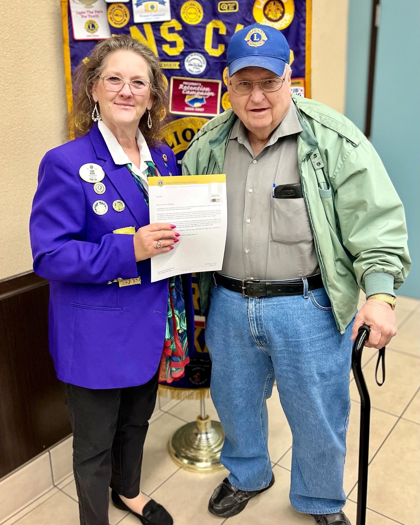 District Gov Vicki Dulin presents Lion Ray with 40 year Chevron award in front of the Sugar Land Lions Club Banner.